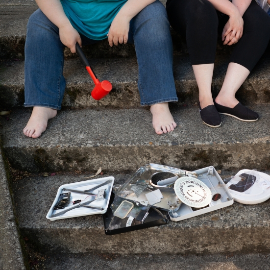 Image is a pile of smashed and broken bathroom scales lies on a concrete step in an urban park. Several women’s feet are resting on the next step up.