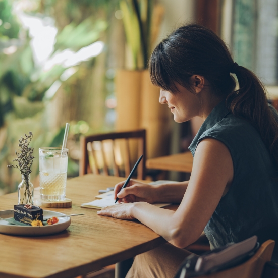 Image of a female sitting at a dining table with a journal and a plate of food She is making notes about something