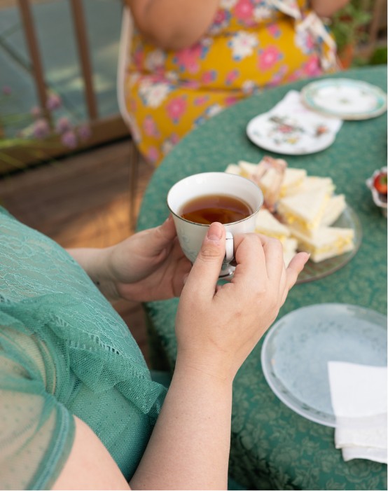 Image is of a plus-size woman enjoying a tea party in a garden on a sunny summer afternoon. She is wearing a mint green dress. On the small table in front of her are a teacup, plate of sandwiches and strawberries