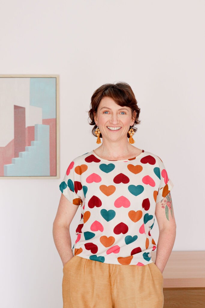 Image of Carly smiling she has brown hair and standing in front of a desk with her hands in pockets and wearing love heart shirt