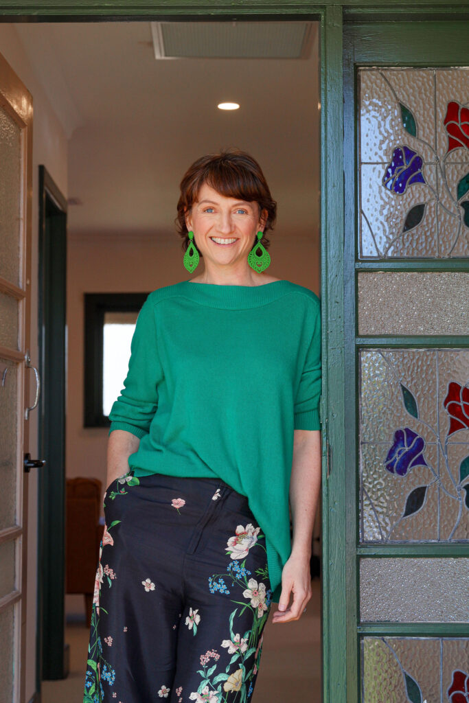 Image of Carly Taylor, she has short brown hair, wearing a green jumper and big green earrings smiling and leaning up against the door frame of her office entrance