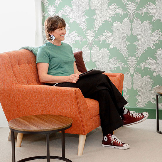 Image of Carly with brown hair sitting in an orange chair with legs crossed smiling at someone out of frame